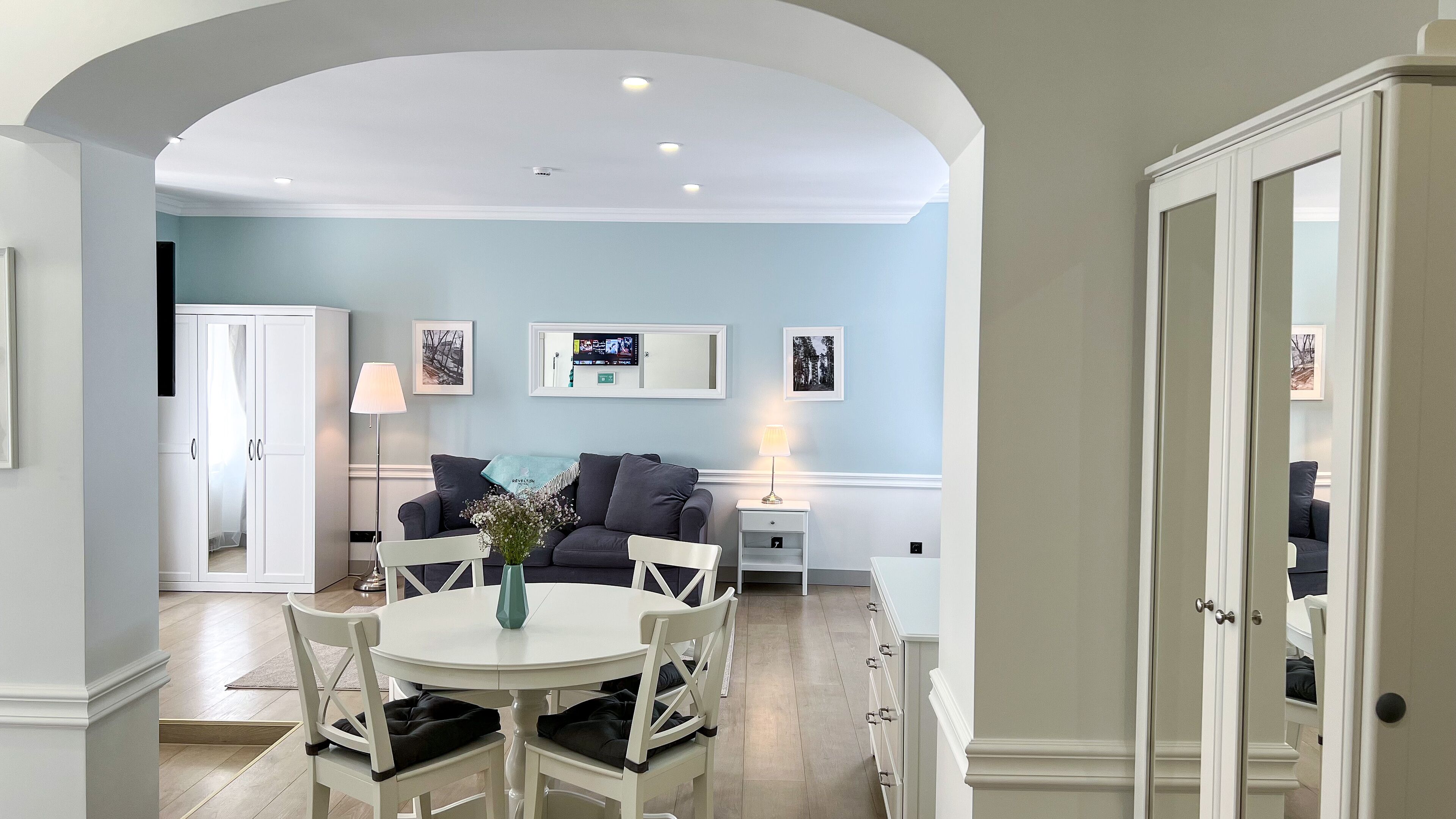 Un salon moderne avec un coin repas, caractérisé par un mur bleu clair, des meubles blancs et un plafond doté d’un éclairage encastré.