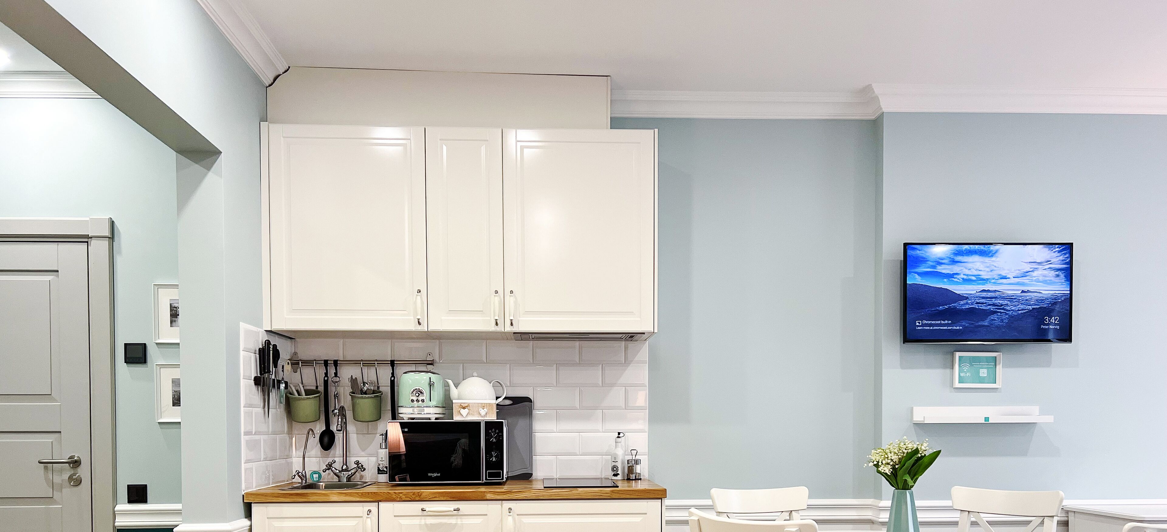 Una cocina moderna con armarios blancos, un microondas y una cafetera. Hay un televisor en la pared que muestra una vista panorámica.