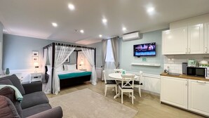 Un dormitorio moderno con una cama con dosel, una zona de estar con una mesa y sillas, una kitchenette con armarios blancos y un televisor de pantalla plana montado en la pared.