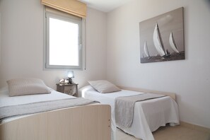A bedroom with two single beds, a nightstand with a lamp, a window with a beige blind, and a sailboat painting on the wall. - Beachfront apartment with sea views - UHC PANORAMIC (Miami Playa)