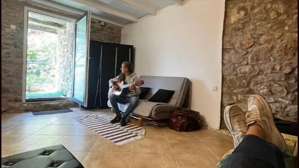 A spacious living area with a stone wall, a sofa, a black ottoman, and a person sitting on the floor. - Refugi per Boletaires i Amants de la Natura (La Nou de Berguedà)