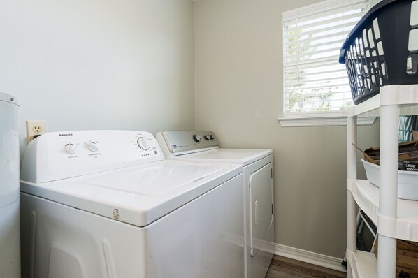 A laundry area with a front-loading washer and dryer, a window with blinds, and a shelf with laundry baskets.