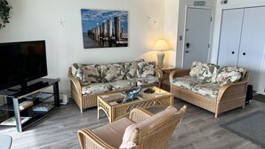 A living room with a sofa, a coffee table, a TV, and a painting of a pier. - Thunder Island 157D-Baywater 39th St, Internet, Pool, Elev, W/D, AC (Ocean City)
