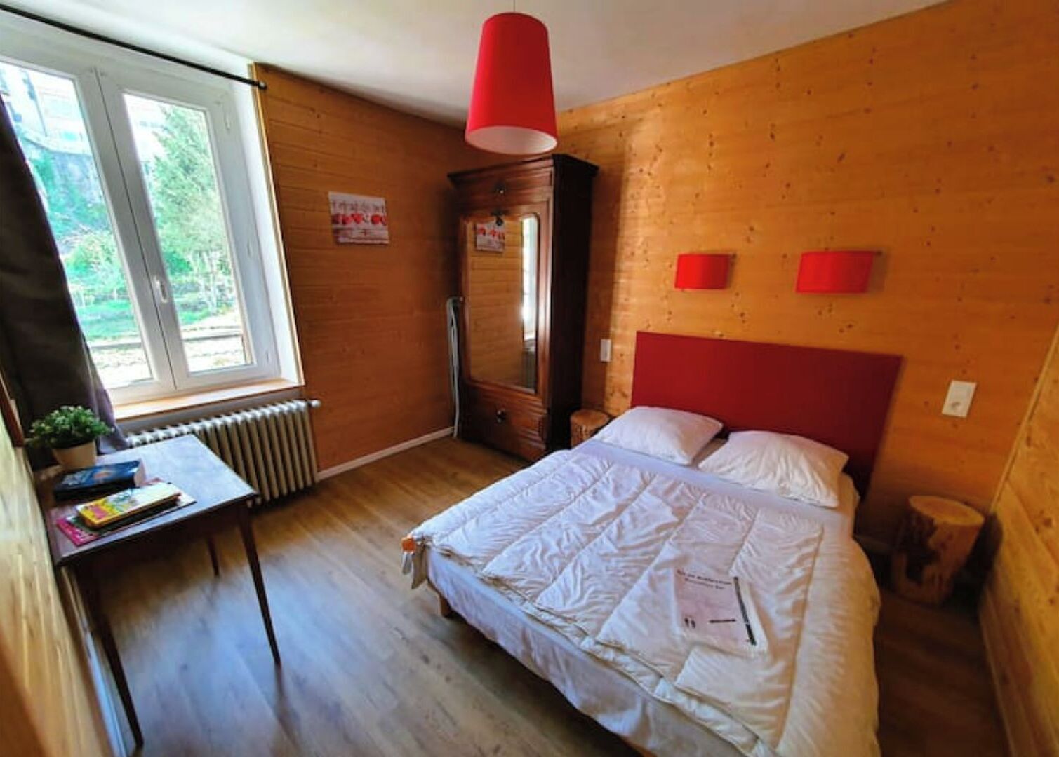 A bedroom with a wooden headboard, a bed with white bedding, a red pendant light, and a window with a view of greenery.