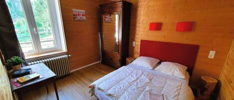 A bedroom with a wooden headboard, a bed with white bedding, a red pendant light, and a window with a view of greenery.