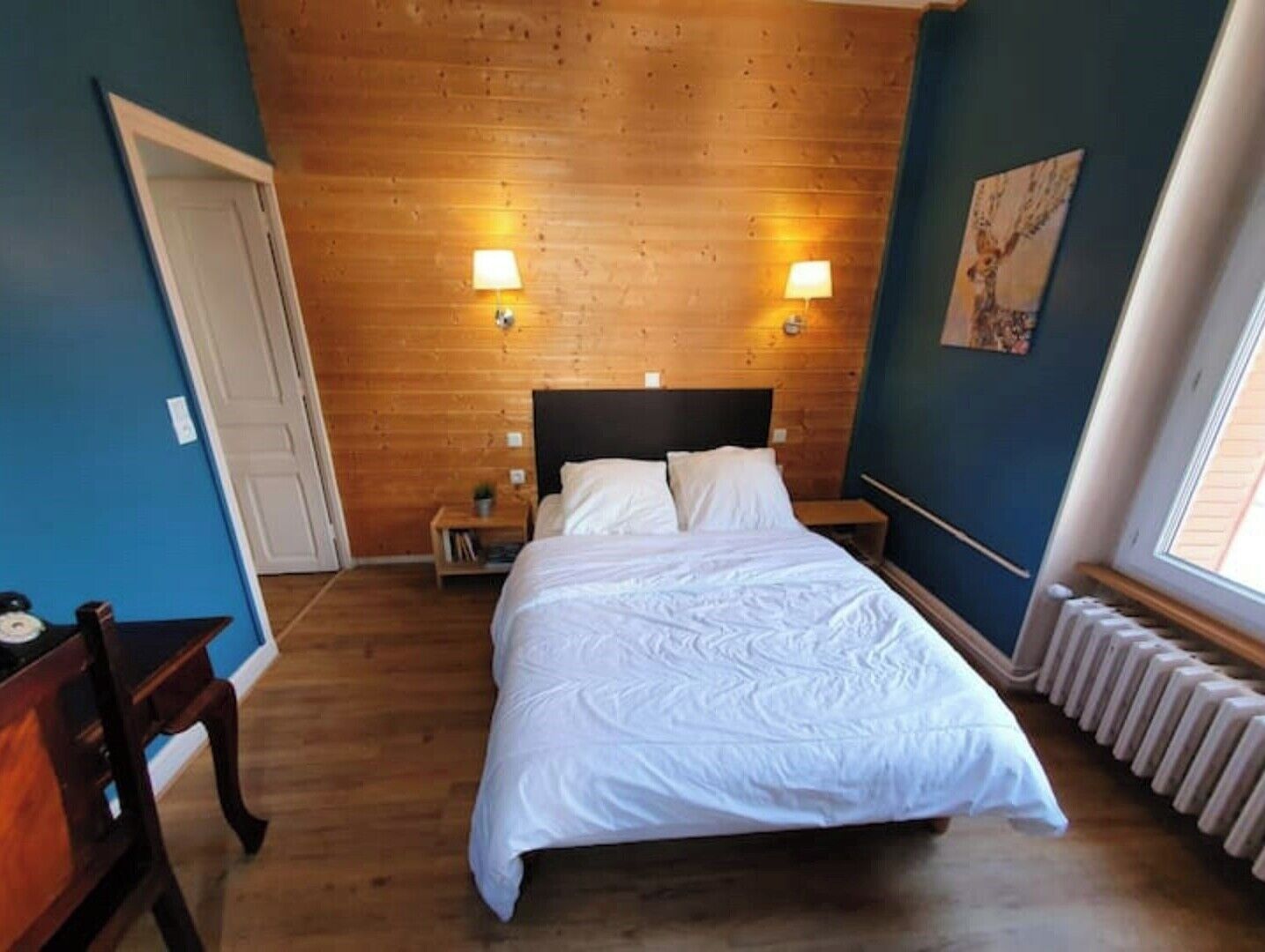 Une chambre à coucher avec une tête de lit en bois, deux lampes de chevet, un lit avec du linge de lit blanc, une petite table de chevet avec une lampe, un radiateur et une fenêtre avec des stores.
