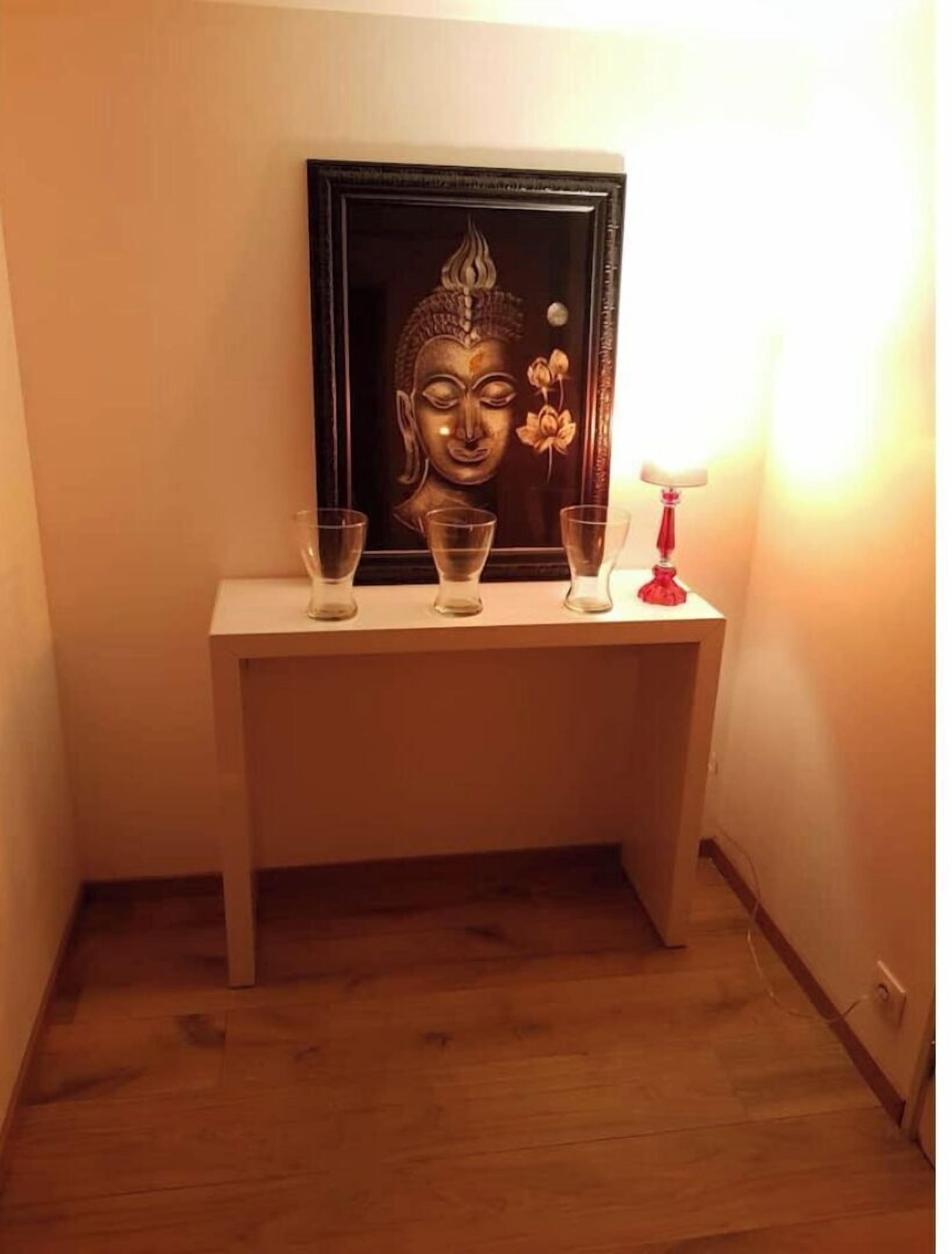 Eine gerahmte Buddha-Statue auf einem weißen Konsolentisch mit drei klaren Gläsern und einer roten Lampe.