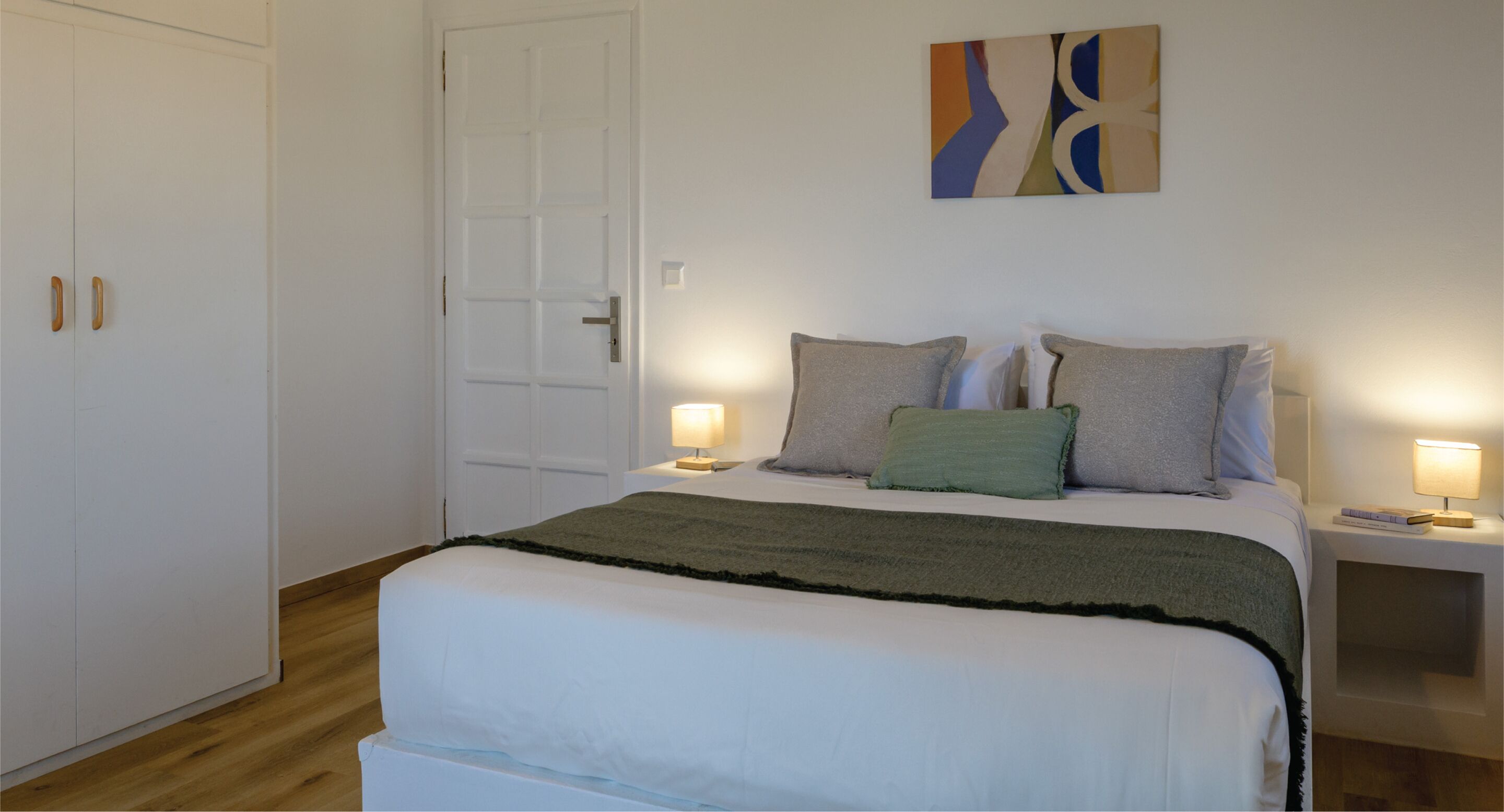 A bedroom with a bed, two bedside tables with lamps, a door, and a wall with a painting.
