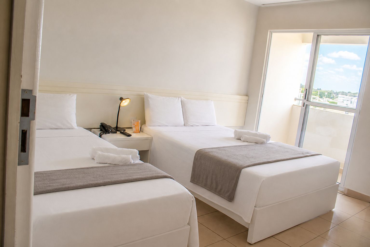Habitación de hotel con dos camas, una mesita de noche con lámpara, teléfono y vistas al exterior.