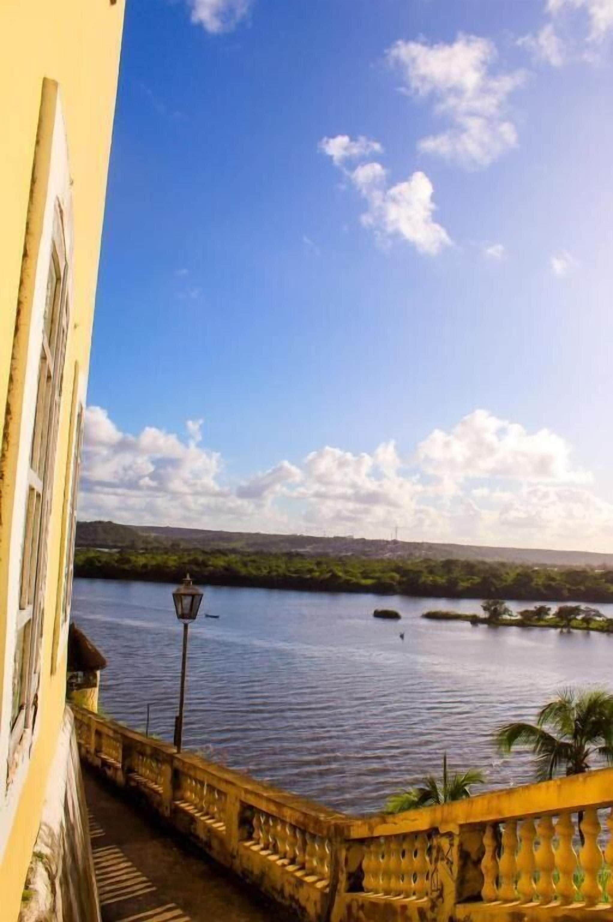 A balcony with a view of a calm body of water, a clear sky, and distant greenery.