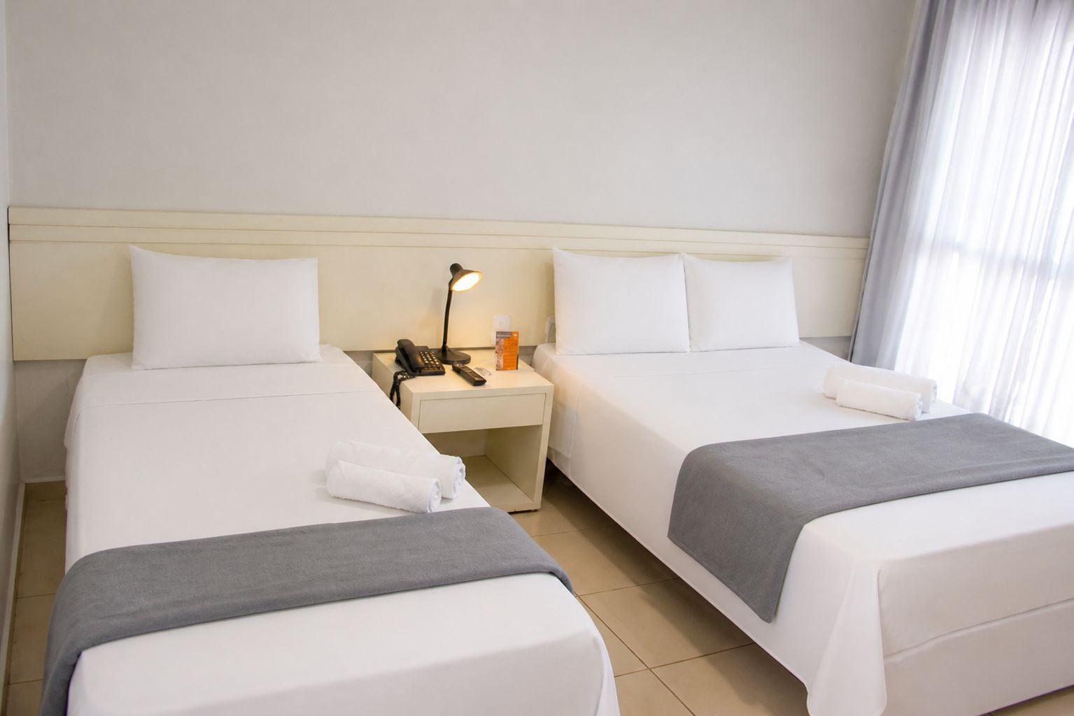 Habitación de hotel con dos camas, una mesita de noche con lámpara, teléfono y ventana con cortinas.