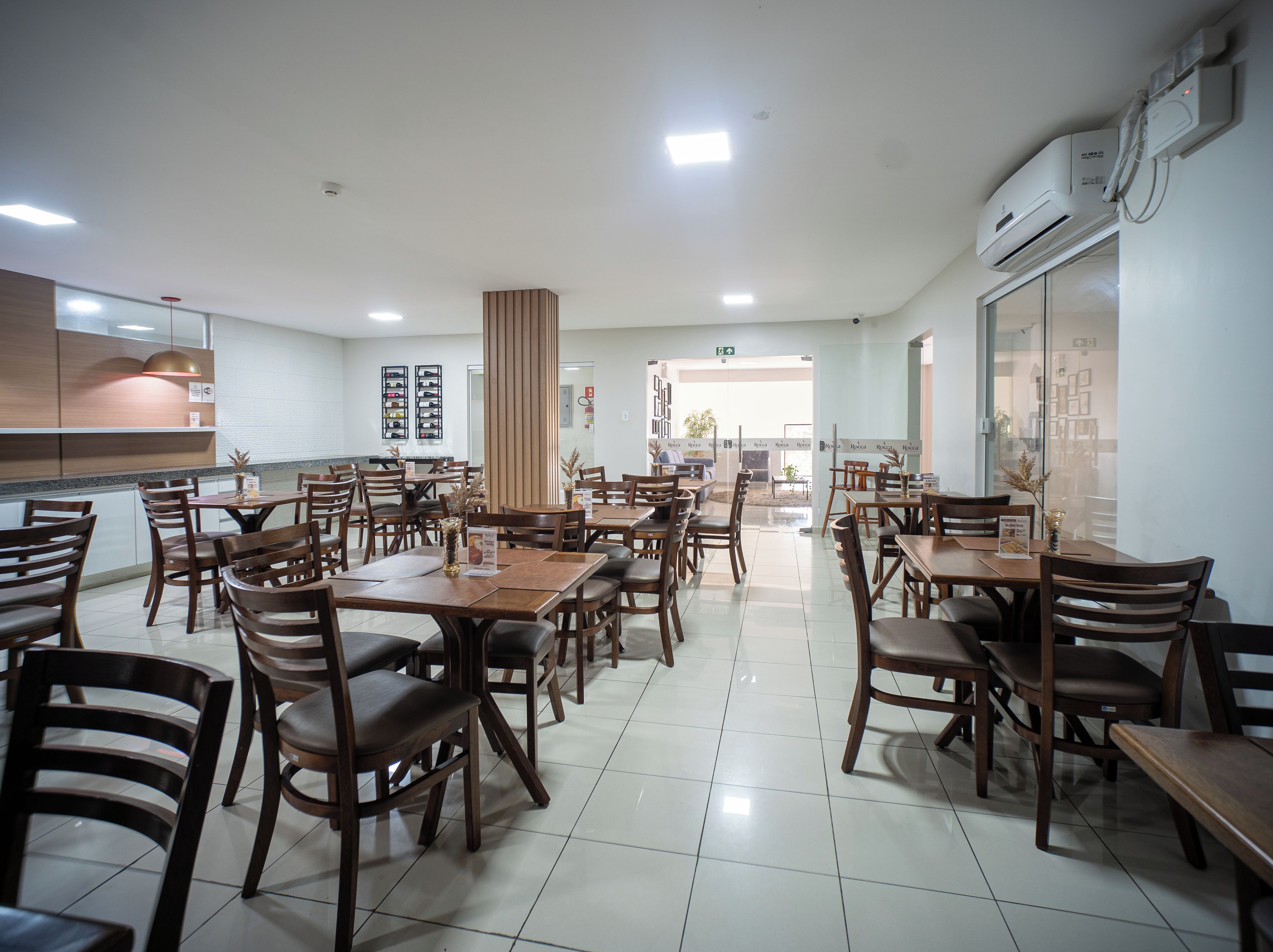 Uma sala de jantar ampla com várias mesas e cadeiras, área de cozinha com balcão e uma janela grande que proporciona luz natural.