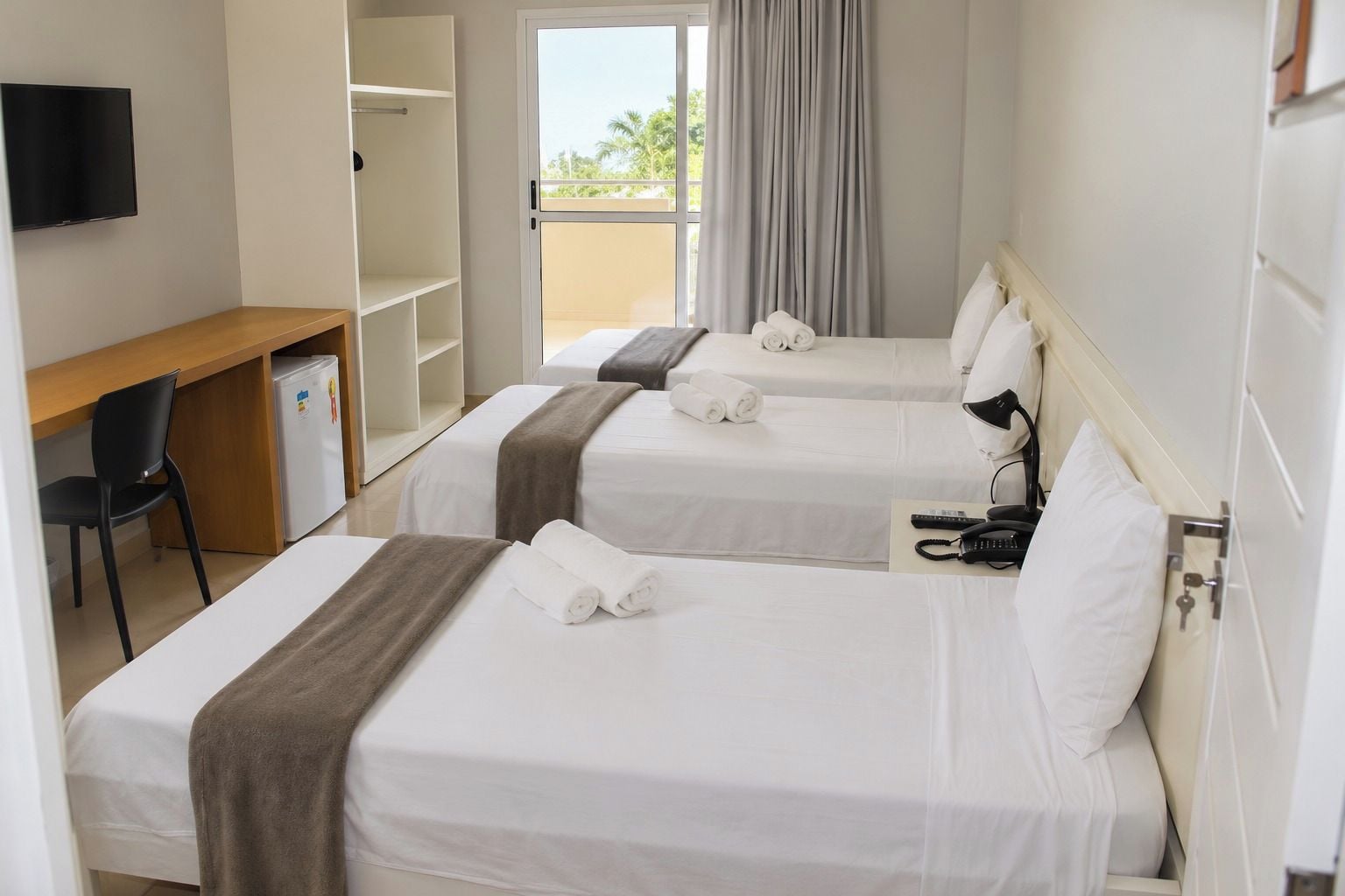 A hotel room with two beds, a desk, a chair, a TV, and a balcony with curtains.