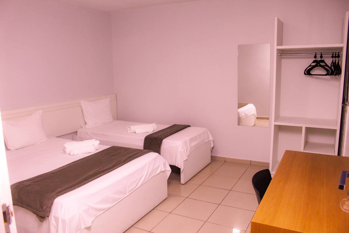 Ein Hotelzimmer mit zwei Betten, einem Holztisch und einem Schrank mit darinliegender Kleidung.