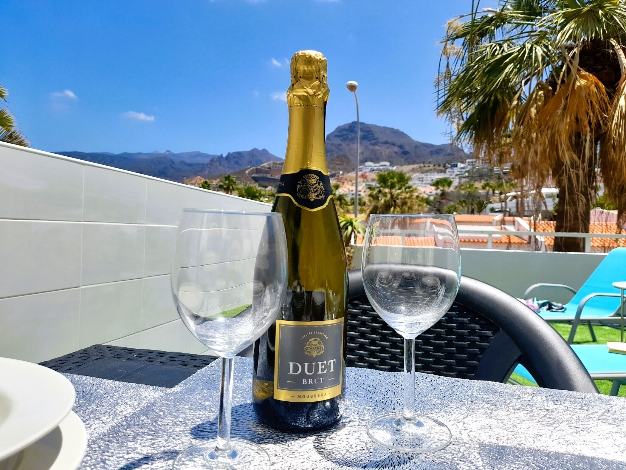 Een fles Duet Brut champagne en twee glazen op een tafel, met uitzicht op bergen en palmbomen.