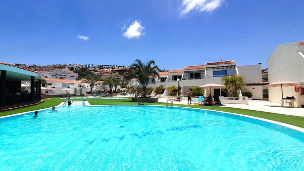 A swimming pool with clear blue water, surrounded by lounge chairs and umbrellas, with buildings and a hill in the background. - Lovely apartment Costa Adeje & Big Terrace (Costa Adeje)