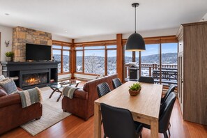 A modern living room with a fireplace, a dining area, and large windows offering a view of a snowy landscape. - Tremblant Prestige - Altitude 172-8 (Mont-Tremblant)