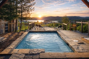 A hot tub with a wooden deck, surrounded by a stone border, and overlooking a scenic view of a lake and mountains at sunset.