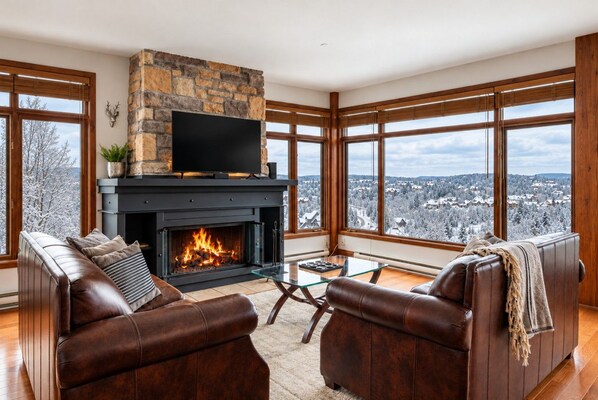 A modern living room with a fireplace, leather sofas, a glass coffee table, and large windows offering a city view.