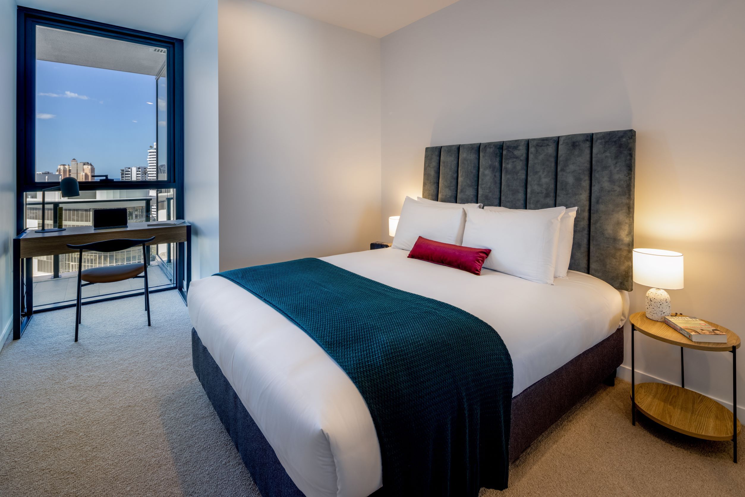 A hotel room with a bed, a desk, a chair, a nightstand, and a view of the city.
