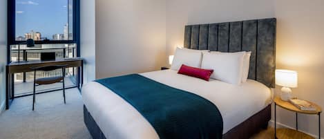 A hotel room with a bed, a desk, a chair, a nightstand, and a view of the city.