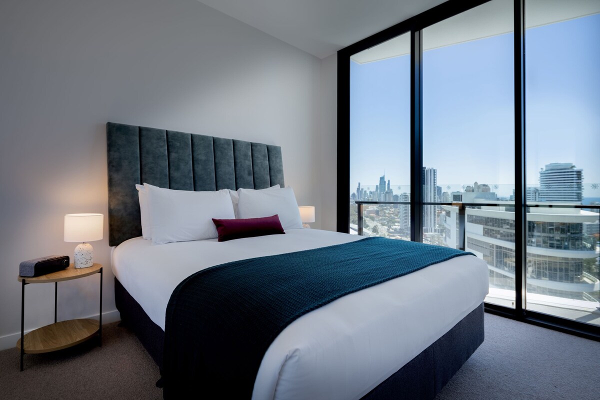 A modern hotel room with a large bed, a nightstand, a lamp, and a city view through large windows.