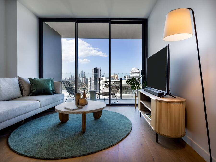 Una sala de estar moderna con un sofá, una mesa de centro, un televisor sobre un mueble bajo y un ventanal con vistas a la ciudad.