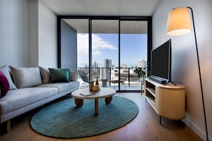 A modern living room with a sofa, a coffee table, a TV on a stand, and a large window with a city view. - The Star Residences Gold Coast (Gold Coast)