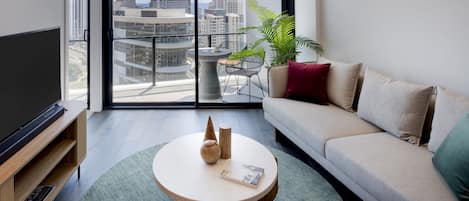 Una sala de estar moderna con un televisor de pantalla plana, una mesa redonda de centro con objetos decorativos, un sofá con cojines y un ventanal con vistas a la ciudad.