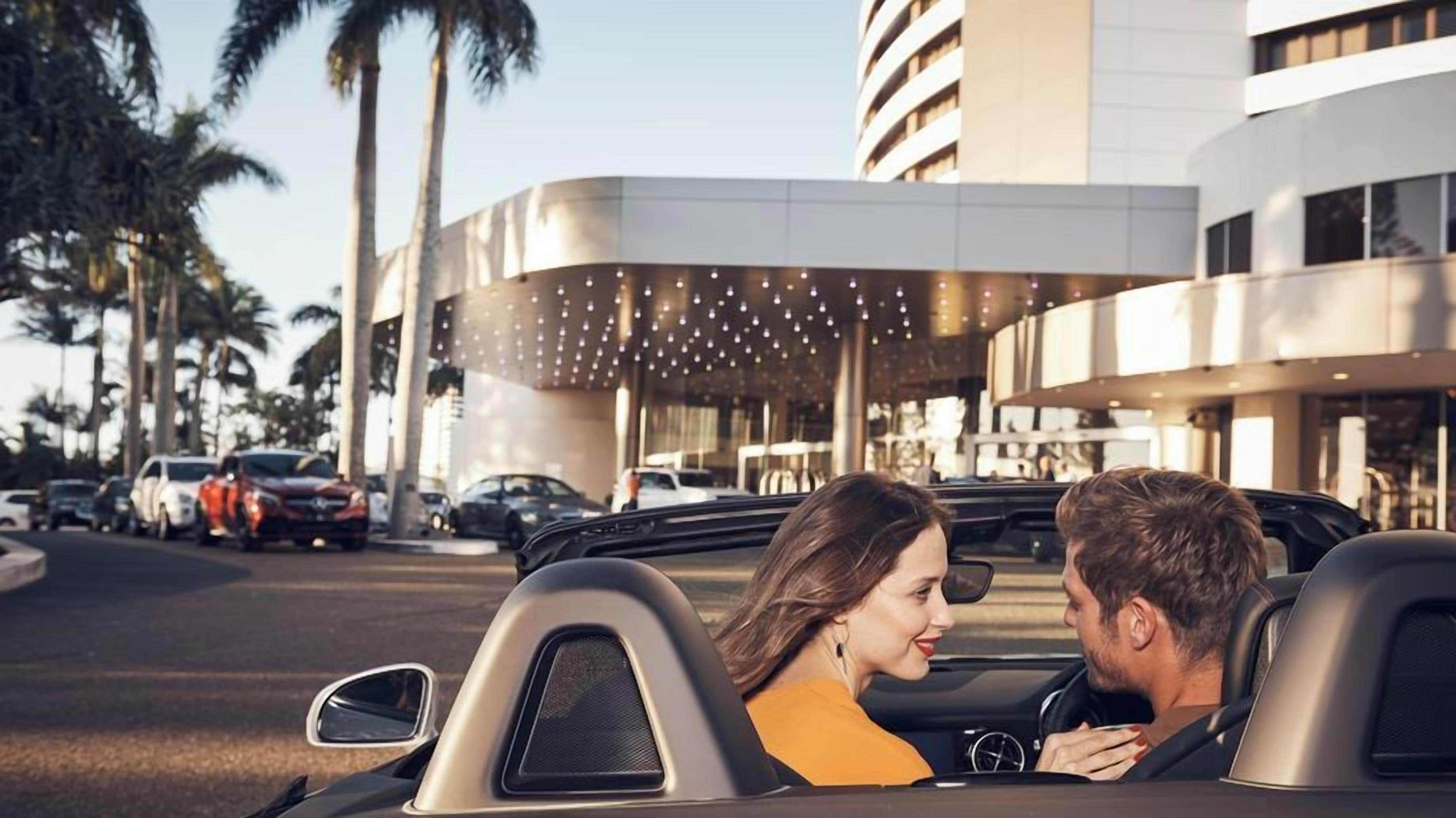 Um carro conversível estacionado em frente a um edifício com design arquitetônico moderno, palmeiras e outros carros estacionados.