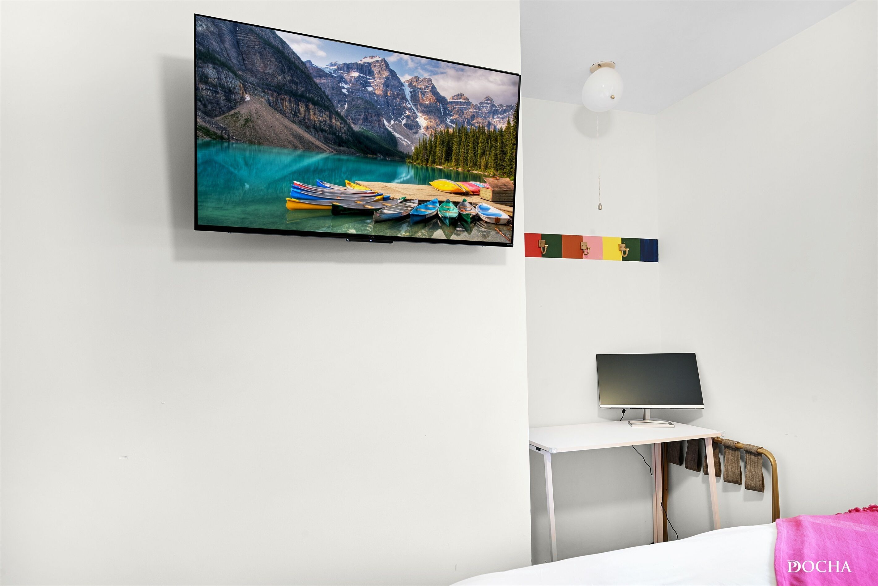 Een slaapkamer met een grote televisie aan de muur waarop een mooi landschap staat, een bureau met een laptop en een bed met een roze deken.