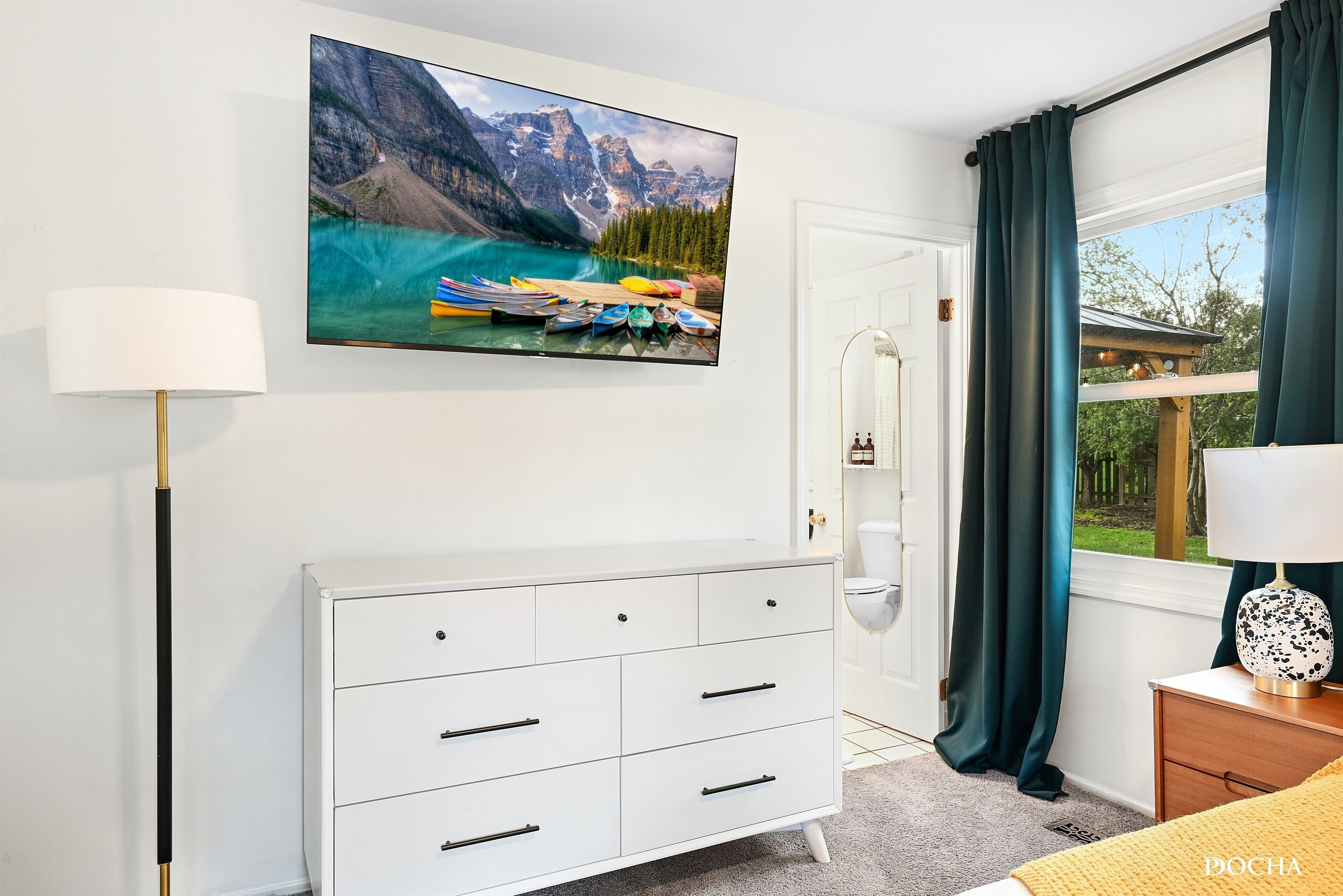A bedroom with a white dresser, a painting of a mountain lake, a bed with a yellow blanket, and a window with curtains.