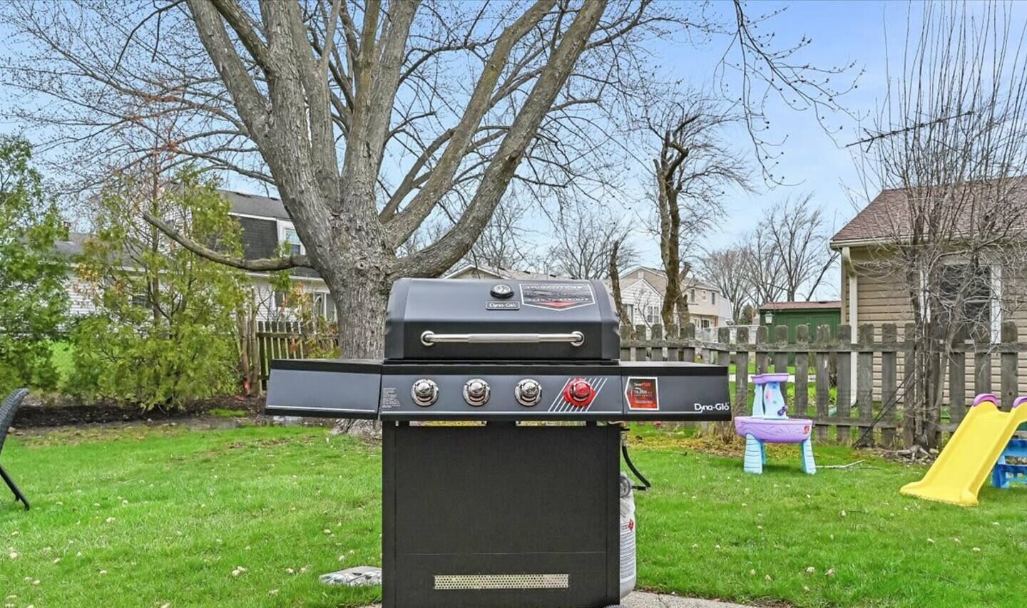 Una parrilla para barbacoa en un patio trasero con valla de madera, un árbol y un parque infantil.