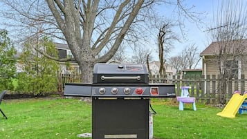A barbecue grill in a backyard with a wooden fence, a tree, and a play structure.