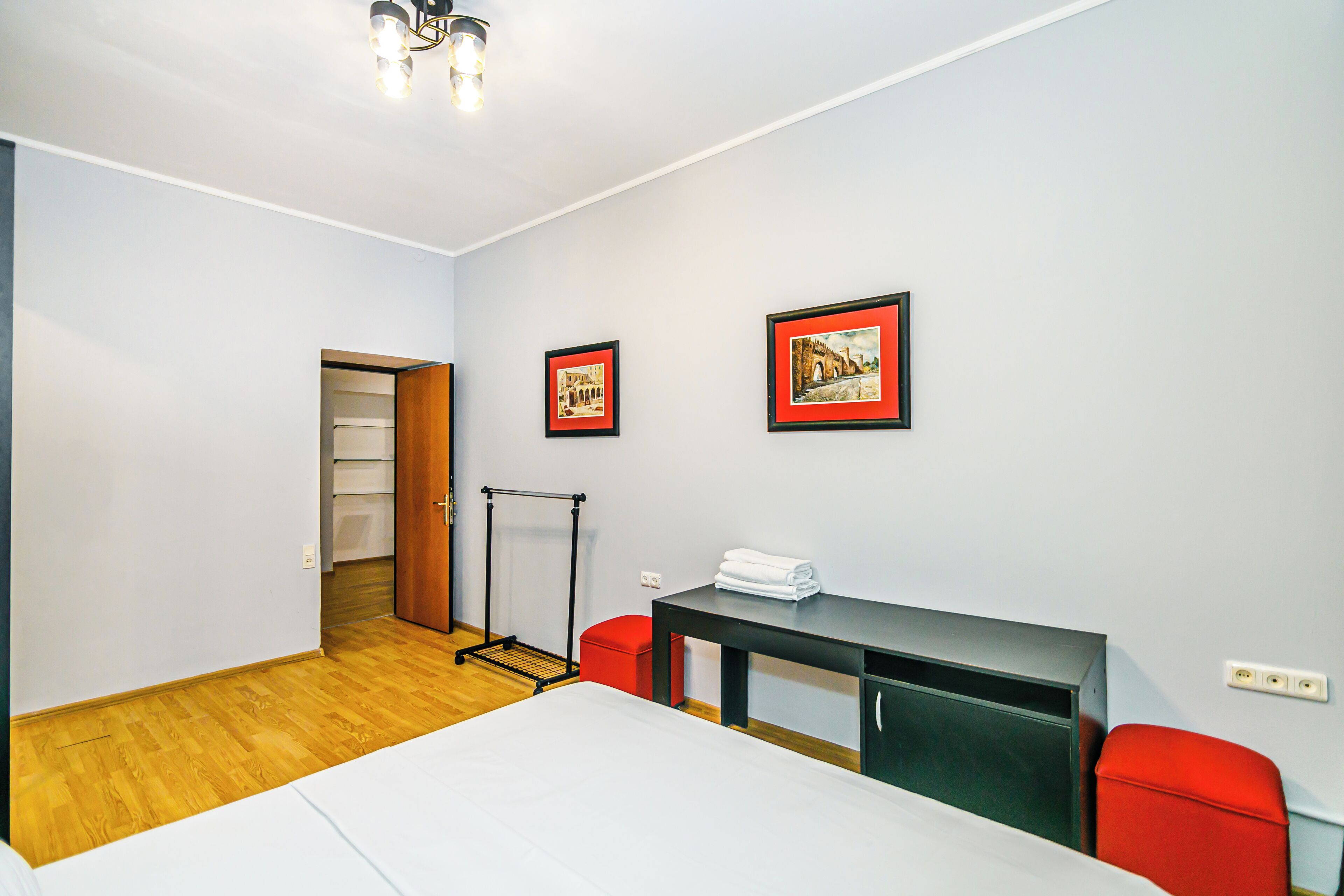 A hotel room with a bed, a desk, two chairs, a red ottoman, a coat rack, and two framed pictures on the wall.