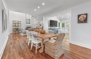A spacious dining area with a wooden table and white chairs, a wicker chair, and a bookshelf with decorative items.