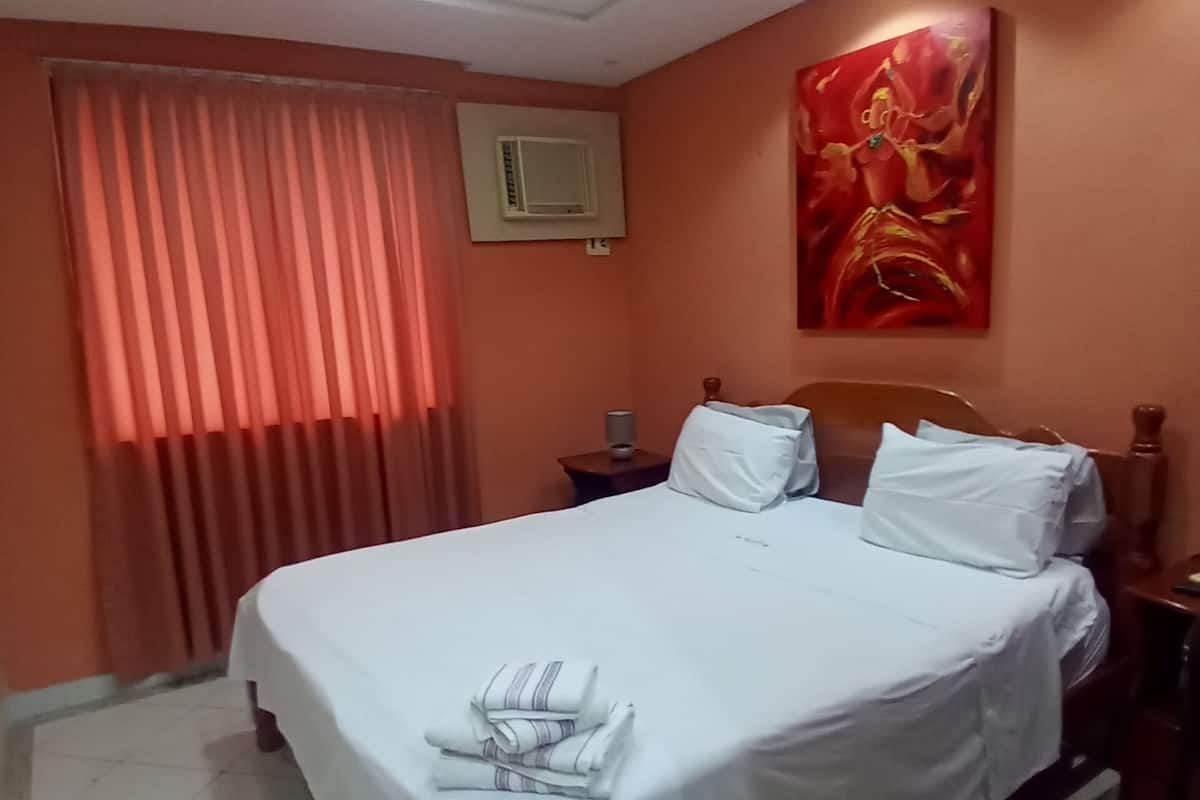 A hotel room with a large bed, a nightstand, a wall-mounted air conditioner, and a window with curtains.