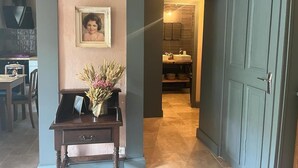 Un couloir avec un bureau en bois, un tableau encadré et un vase de fleurs. Une porte donne sur une salle de bain équipée d’un lavabo et d’un miroir.