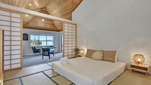 Une chambre de style japonais traditionnel, avec un sol recouvert de tatamis, un lit avec une literie blanche, un bureau avec une chaise et une vue à travers un paravent shoji.
