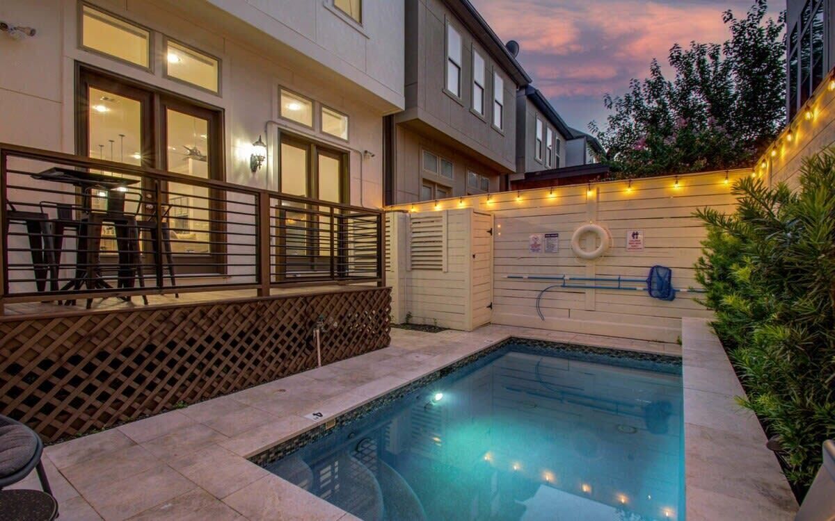 A swimming pool with a wooden deck and a fence, surrounded by buildings with windows and a sign.