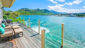 Eine Holzterrasse mit Gartenmöbeln mit Blick auf das klare, blaue Meer und üppige, grüne Hügel.