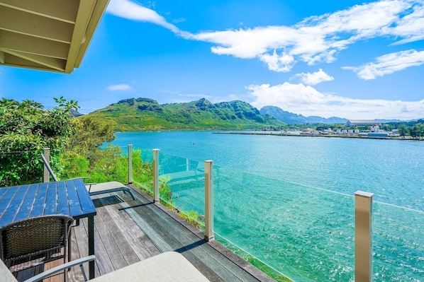 A wooden deck with a glass barrier overlooking a clear blue lake, surrounded by greenery and mountains.