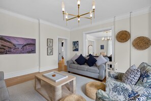 A living room with a grey sofa, a coffee table, and a patterned armchair. There is a painting on the wall and a chandelier above the sofa.