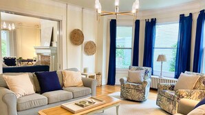 A living room with a sofa, armchairs, a coffee table, a fireplace, and a large window with curtains. - Tazewell House Estate with Wraparound Porch (CAPE CHARLES)