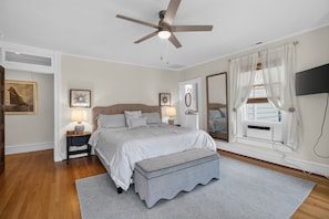 A bedroom with a bed, bedside tables, a ceiling fan, a window with curtains, a mirror, and artwork on the walls.