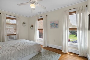 Une chambre à coucher avec un lit, un ventilateur de plafond, une fenêtre avec des rideaux, un tableau et une vue sur l’extérieur.
