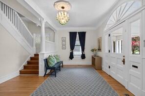 A hallway with a staircase, a bench with cushions, a patterned rug, a chandelier, and a door with a window view.