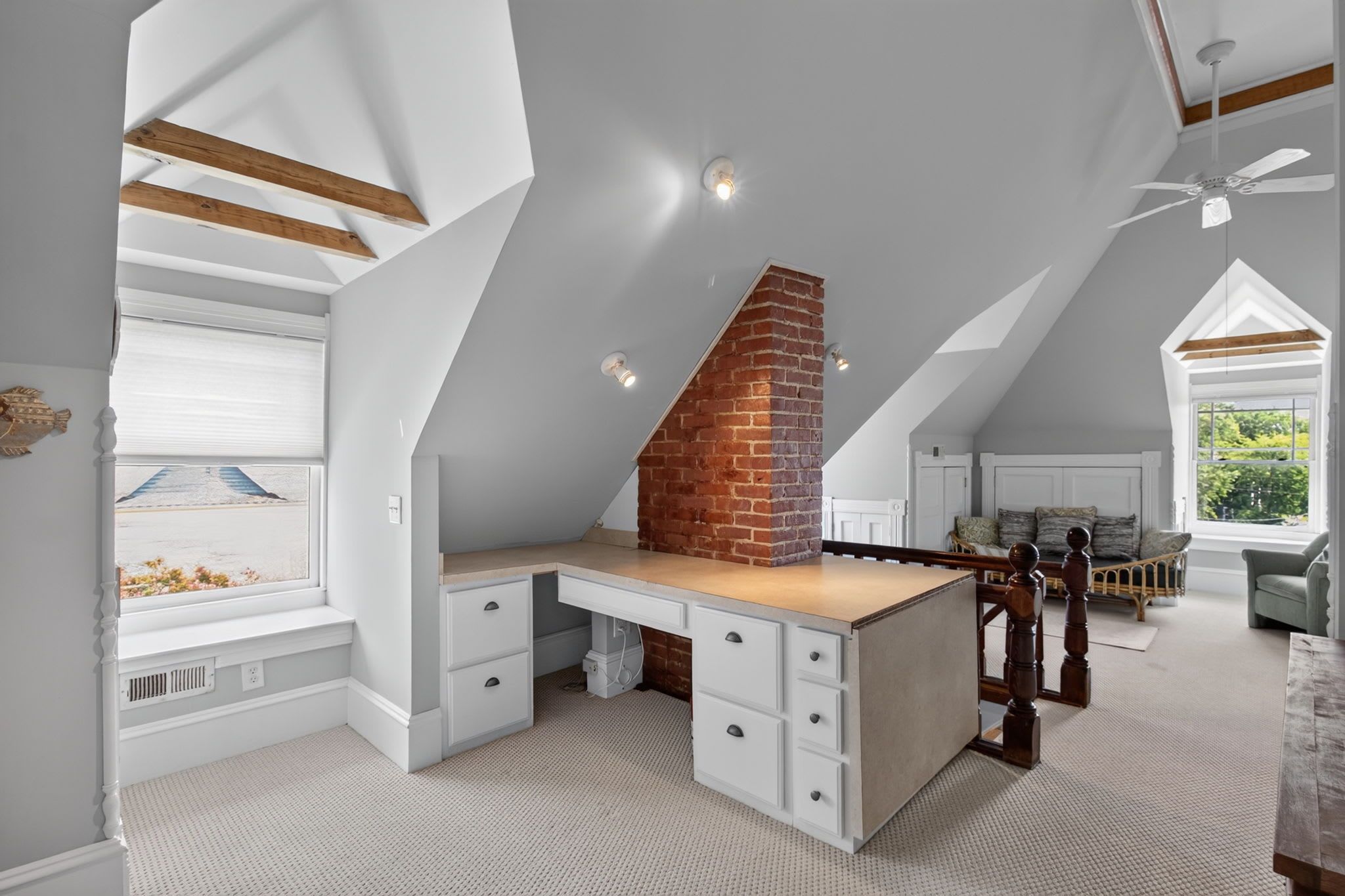 A loft bedroom with a sloped ceiling, a brick fireplace, a desk with drawers, and a view of a beach through a window.