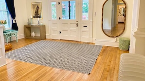 A room with a wooden floor, a patterned rug, a white door with glass panels, a round mirror, and a small table with a vase. - Tazewell House Estate with Wraparound Porch (CAPE CHARLES)