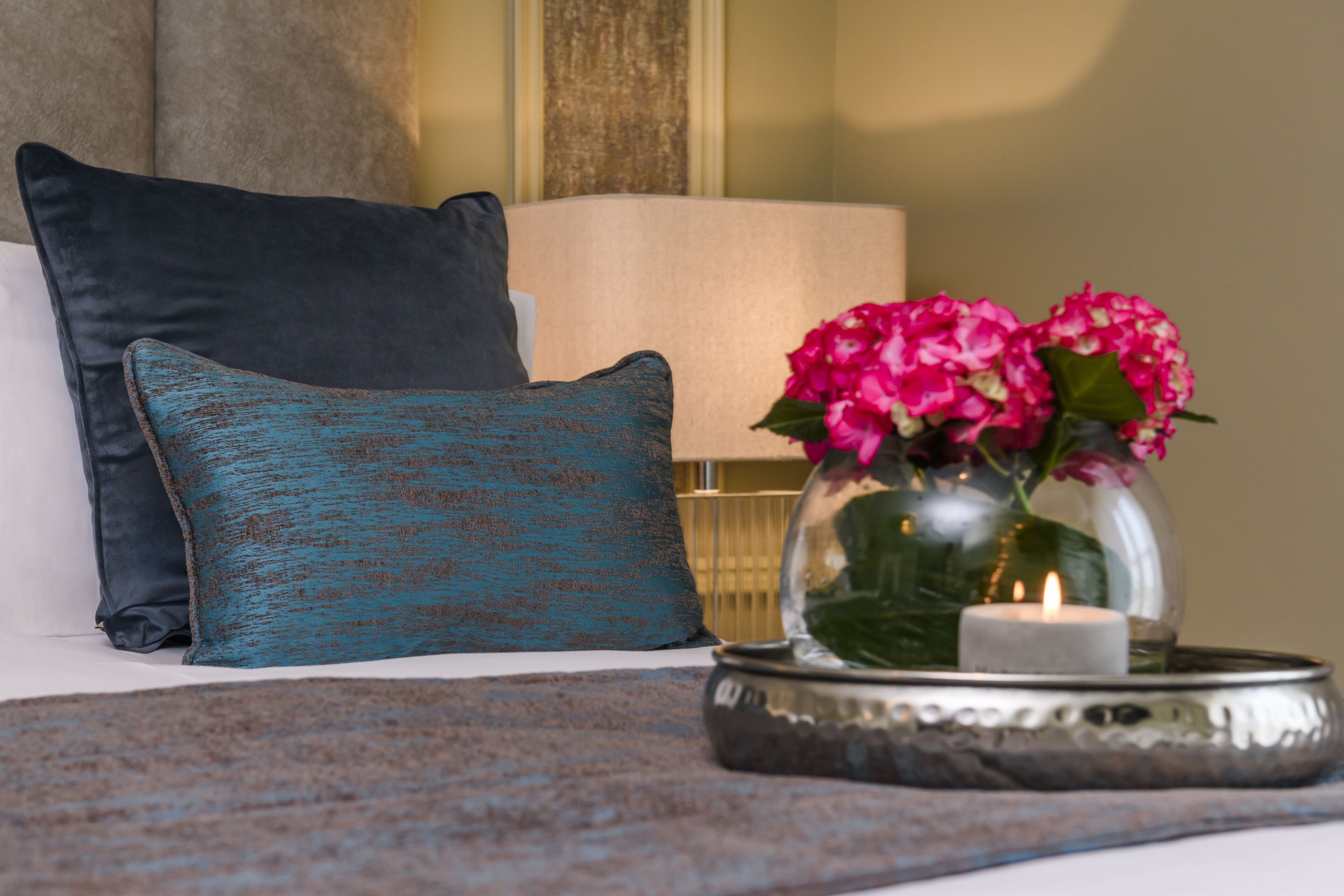 A neatly arranged bed with a teal pillow, a white pillow, and a decorative teal pillow with a distressed finish. A glass vase with pink flowers and a lit candle are placed on a metallic tray beside the bed.